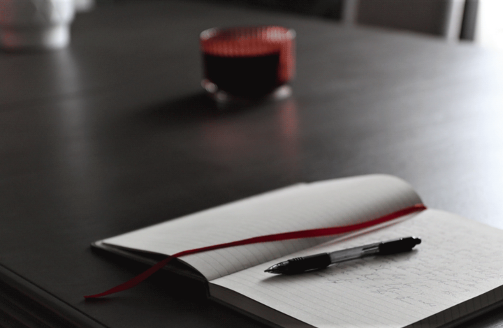 Open journal with a pen next to a red ribbon divider on a grey table. The open pages filled with goals to achieve for the year.